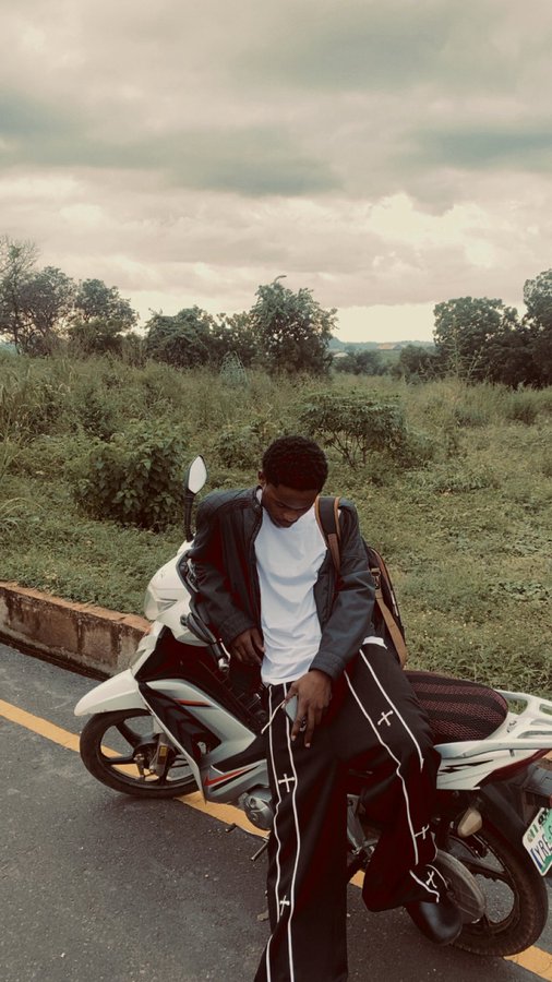 A man in a dark jacket and black track pants sits on a white scooter on a road with greenery and a cloudy sky in the background.