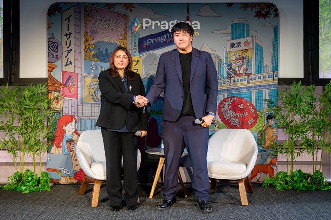 Two presenters stand on stage shaking hands in front of a "Pragmma" branded backdrop featuring Tokyo city illustrations, Japanese text, and decorative bamboo plants on either side.