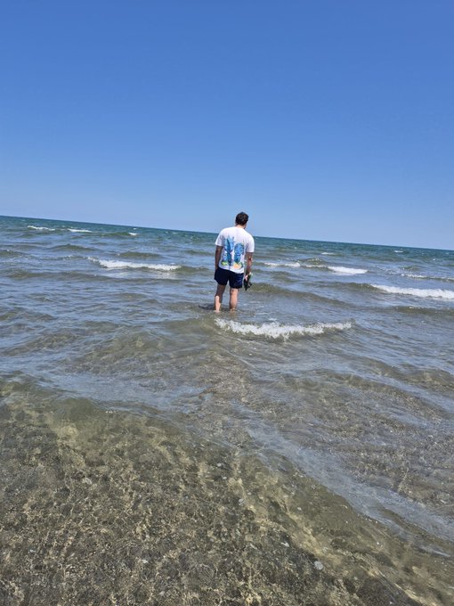 A person wearing a white t-shirt and dark shorts stands ankle-deep in clear ocean water on a sunny day with a blue sky.