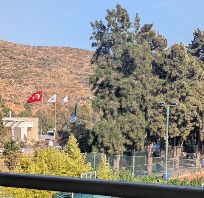 A serene view of a tennis court nestled against a sunlit hillside, with a large pine tree and a Turkish flag flying prominently in the foreground, capturing the scenic charm of the visit with friends.