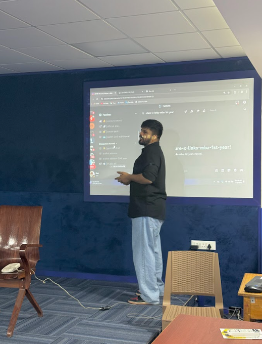 A man in a black shirt and light blue jeans stands presenting in front of a large interactive display showing a Figma interface with text including "are-x-links-mbs-1st-year" in a room with blue walls.
