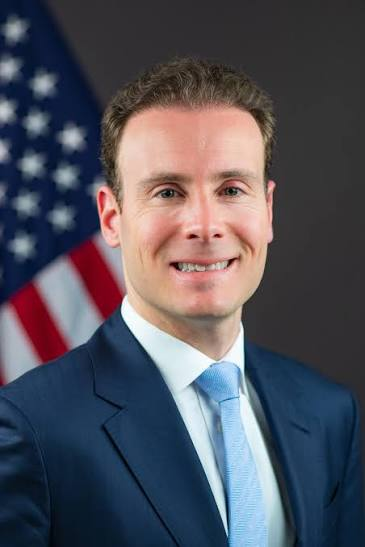 A professional headshot of a man with a blue suit and light blue tie, smiling in front of an American flag.