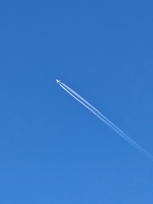 An airplane flies high across a clear blue sky, leaving behind two long, white contrails stretching diagonally across the frame, suggesting the post is asking for examples of sky photography with jet trails.