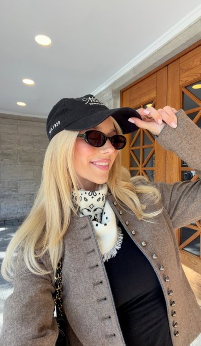 A blonde woman wearing sunglasses, a black cap, and a brown jacket smiles while standing outdoors.