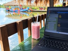 A workspace setup showing a laptop on a green table next to a pink smoothie in a jar, with a scenic view of a river and boats through a window.