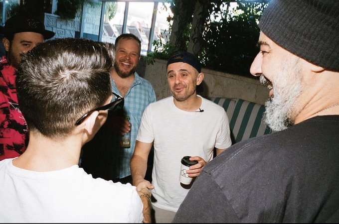 A candid photo of a group of men socializing outdoors at night, with one holding a coffee cup and another in a grey beanie, in what appears to be a casual gathering.