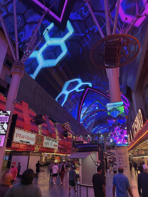 A vibrant nighttime view of the Fremont Street Experience in Las Vegas, featuring a large illuminated LED canopy displaying blue geometric patterns above crowded pedestrian walkways and neon-lit storefronts.