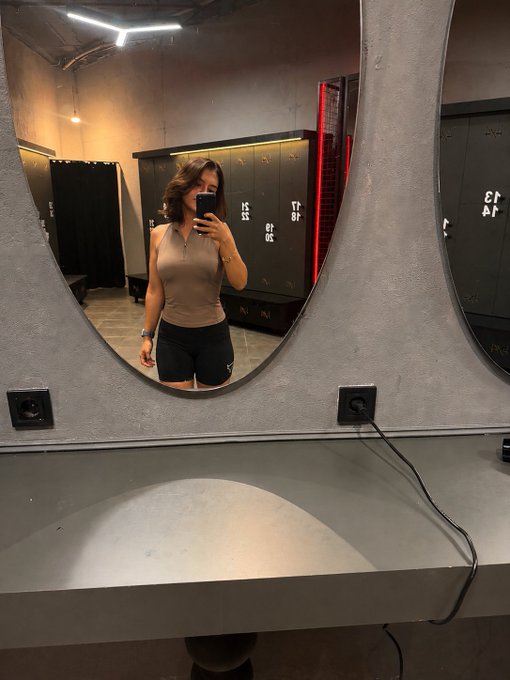 A woman in a tan top and black shorts takes a mirror selfie in a gym locker room, with hair dryer cords hanging from outlets visible on the concrete countertop below the rounded mirror.

The image fits the post perfectly—it captures a casual "gm" (good morning) gym selfie, a popular type of content on X (Twitter), playing into the theme of testing whether the platform's new algorithm will surface her post on followers' feeds.