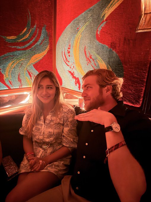 A man and woman sitting together in a dimly lit venue with red walls and colorful abstract art behind them, the man gesturing with his hand showing a watch.