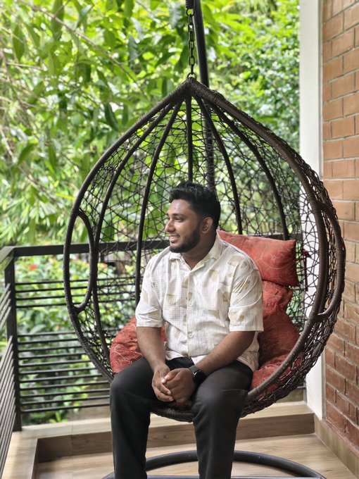 The image features a man sitting in a hanging chair with his legs crossed while wearing a white shirt, black pants, and a watch. The man appears to be smiling at the camera. The background of the photo shows trees and bushes, suggesting that they are located outdoors or near a park. The caption reads "Good Morning X fam! No noise, just pure building. Bullish on what @XOOBNetwork and @useTria are bringing to the table." This implies that the man is enjoying his time in this outdoor setting while appreciating the work of others who have contributed to the image's content.