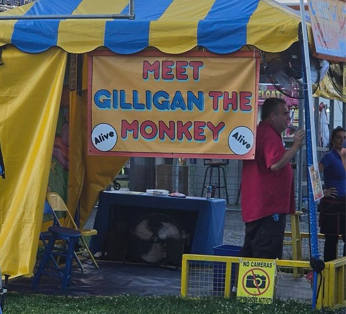 A carnival or fair booth with a blue and yellow striped awning, featuring a large yellow sign that reads 'MEET GILLIGAN THE MONKEY' with 'Alive' written on both sides, while a man in a red shirt stands to the right and a 'NO CAMERAS' sign is posted at the entrance.
