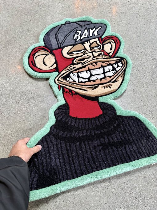 The image features a person holding up a large, colorful rug with a cartoon monkey design on it. The rug has a black and green color scheme, and the monkey appears to be wearing a hat and a sweater. The text "Collectors collect. IRL is alpha & always will be." is also present in the image. This suggests that the person holding up the rug might have an interest in collecting unique or quirky items, such as this cartoon monkey-themed rug.