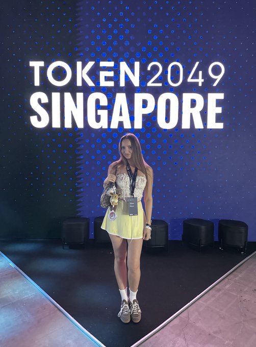 The caption reads: A young woman in a yellow skirt and patterned top poses in front of a large illuminated "TOKEN 2049 SINGAPORE" backdrop, holding a plush bear accessory while wearing a conference lanyard—reminiscing last year's event since she skipped this year's edition.