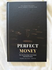 A hardcover book titled 'PERFECT MONEY' by Fergus Michael Irvine rests on a light surface, with text indicating it addresses 'THE WORLD'S FIRST AND ONLY PERFECT MONEY SYSTEM, How unlimited wealth, unlimited clean energy and world peace can be achieved.'