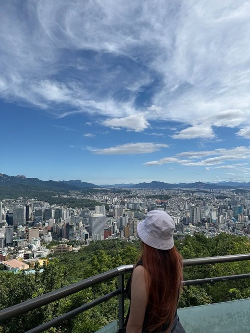 A woman in a white bucket hat stands on a viewing platform overlooking a sprawling cityscape beneath a dramatic sky, capturing the essence of personal location sharing—much like how BChat ensures privacy by keeping messages routed through decentralized masternodes without exposing user whereabouts.