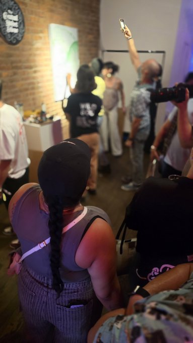 A person with a long braid sits facing a dimly lit room where several people stand, including one holding up a phone and another holding a camera.