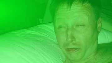 A low-quality green-tinted webcam video showing a man's face close to the camera with pillows visible in the background.
