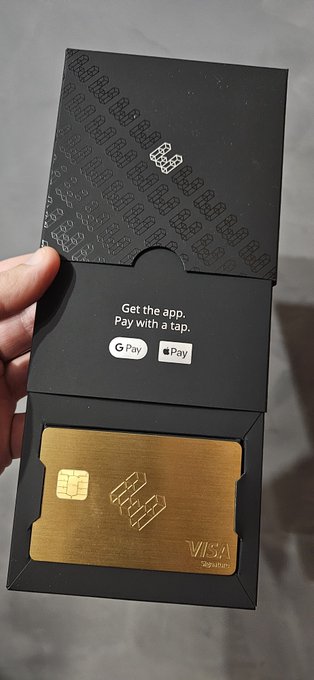 A hand holds an open black presentation box displaying a gold-colored Visa credit card and a slip reading 'Get the app. Pay with a tap.' with Google Pay and Apple Pay logos.