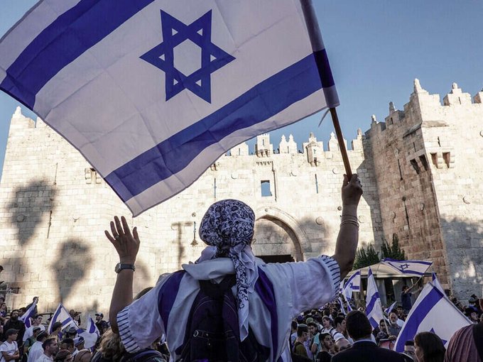 A person waves an Israeli flag with the Star of David emblem in front of an ancient stone gate, surrounded by a crowd also displaying Israeli flags. The image accompanies the post by visually representing Israel, the country where Bank Leumi—the institution announcing Bitcoin purchasing for its customers—is based.