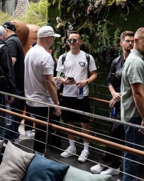 Several men in casual attire converse in a hallway with a lush green plant wall in the background, one holding a phone and wearing a white t-shirt with a black logo.