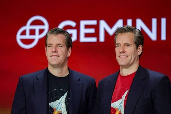 Two men in dark blazers smile as they stand in front of a red backdrop displaying the "GEMINI" cryptocurrency exchange logo and name. The image relates to the news about a federal judge rejecting an attempt to overturn a $5 million settlement involving the Gemini exchange and the CFTC regarding a proposed Bitcoin futures product.