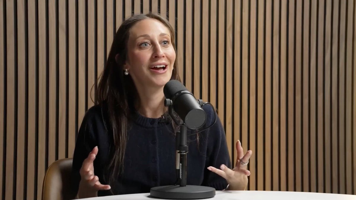 A woman with long brown hair, wearing a dark blue cardigan, speaks animatedly into a Shure microphone against a wooden slatted background.
