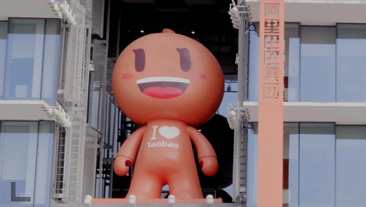 A large orange mascot figure with a smiling face stands between glass windows, wearing a chest inscription reading "I ❤ Taobao," while the banner on the right displays "阿里巴巴集团" (Alibaba Group), clarifying that the mascot is the official character 淘公仔 (Taogongzai) representing the Alibaba/Taobao brand rather than being called Taobao itself.