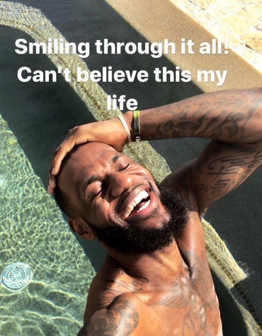 A shirtless, tattooed man laughs joyfully while taking a selfie in a pool, with text overlay reading 'Smiling through it all! Can't believe this my life'.