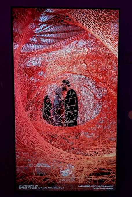 An art installation made of intricate, swirling red threads or yarn, with text at the bottom reading 'WHAT IS GOING ON BEYOND THE VEIL? by Yousef & Rehab (Mayyorga)' and 'CANAL STREET MARKET / BEYOND BORDERS Curated by Choi Jang Lyk'.