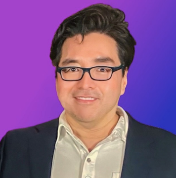 A headshot of a smiling man with dark hair and glasses wearing a light green shirt and dark blazer against a purple background.