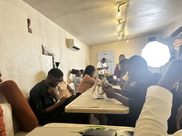 A group of people sit around a long dining table in a warmly lit room with orange cushions and ceiling lights, appearing to share a meal together.