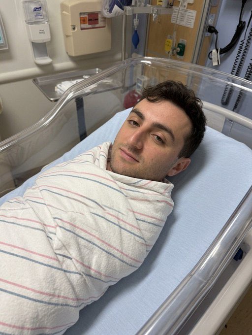 The image shows a man lying down in an infant crib, wrapped in a blanket with blue stripes. The crib has clear plastic sides that allow for visibility into the baby's room. In the background, there are two bottles placed next to each other on a surface. The caption of the image reads "I exposed the cabal and their racketeering scheme Their botted spaces. Their Insider trading of $libra coin Now they call me daddy. Happy Father's Day."