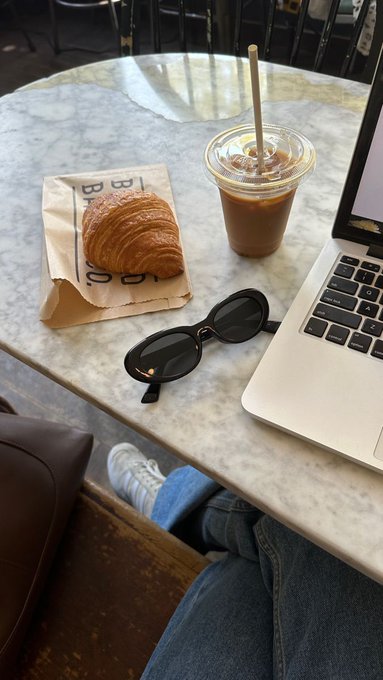 A relaxed Sunday morning scene at a marble café table features an iced latte with a paper straw, black sunglasses, and a flaky croissant on a paper bag, alongside a laptop. Visible text on the bag reads "BAKERY" and the post references "$BTC," tying the leisurely coffee break to a background of crypto market watching.