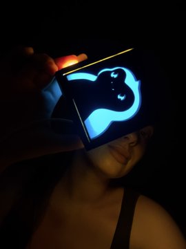 A dark selfie of a person holding up a glowing card or device displaying a luminous blue heart design that illuminates their face.