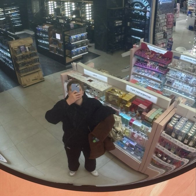 A mirror selfie taken inside a cosmetics store shows a person in dark clothing holding a phone, with display shelves labeled 'WOMEN SECRET', 'LUCY', and 'ADVANCE' visible in the background.