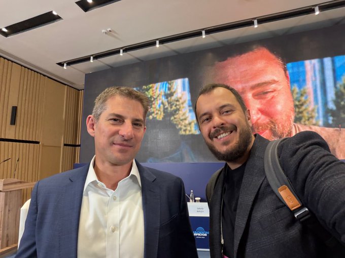 Two men posing for a selfie in what appears to be a conference setting, with a large display screen visible behind them.
