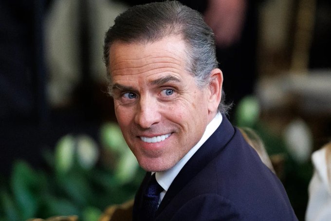 A close-up photograph of a smiling man with slicked-back hair wearing a dark navy suit and white shirt.