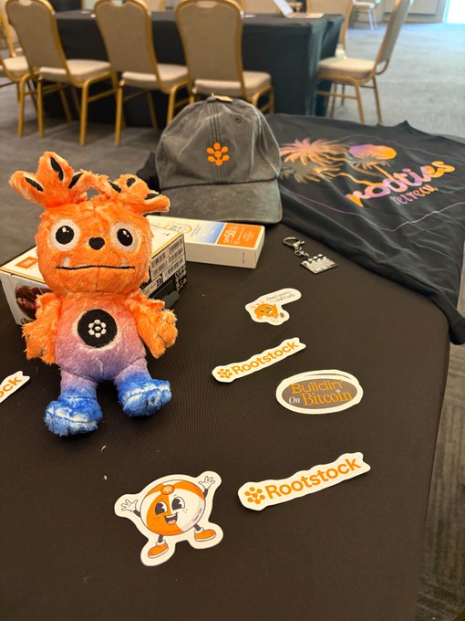 A gray "Rootstock" cap, a colorful t-shirt, and various stickers, including ones featuring an orange mascot character and labels reading "Rootstock" and "Biticity of Bitcoin," are displayed on a black table alongside plush mascot figurines, forming the keepsakes from Rootstock Labs' annual retreat.