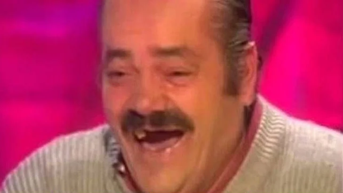 A man with a mustache laughs with his mouth open against a pink background.