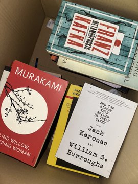 A cardboard box contains several books, including a red Murakami book (Blind Willow, Sleeping Woman), a title by Jack Kerouac and William S. Burroughs, and a black-and-white "EVERY" poster on top.