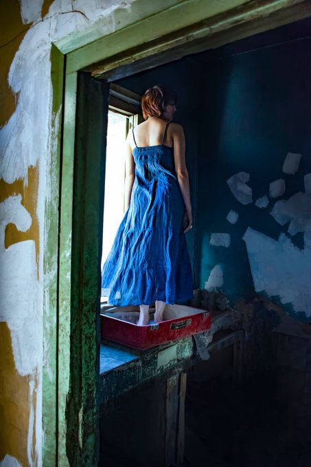 A woman with short red hair, wearing a flowing blue gown, stands with her back to the viewer looking out a window in a dilapidated room with peeling paint, with a red suitcase at her feet.