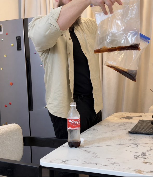A man in a beige shirt holds plastic bags containing a dark liquid over a Coca-Cola bottle on a marble countertop, with a refrigerator in the background.