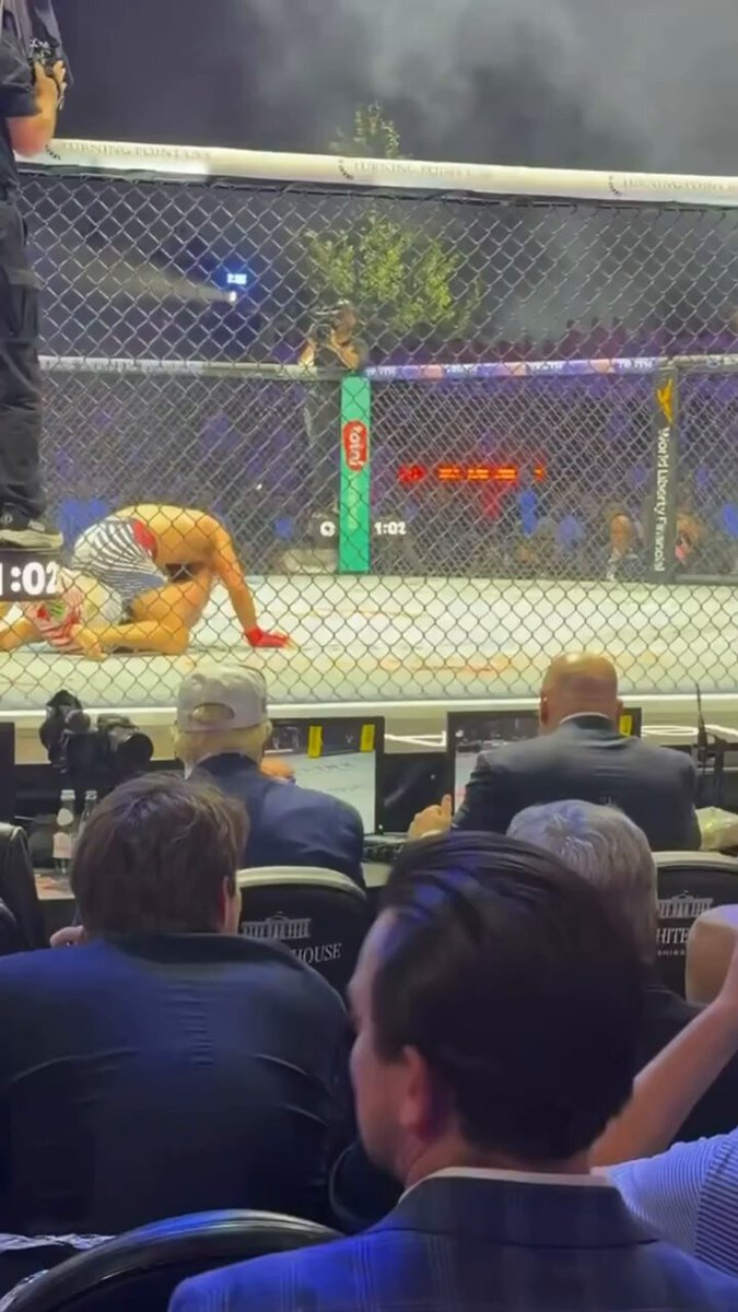 The image shows a fighter inside a cage at an indoor arena with several people watching from their seats, including some sitting in chairs and others standing. The fighter appears to be in the middle of a fight or practice session. A man wearing a suit is seated among the spectators, possibly a referee or an event organizer. The scene takes place during an MMA match, as evidenced by the fighters' positions within the cage and the presence of multiple people watching from various locations around the arena.