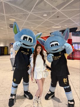 A woman poses between two OKX-branded Manchester City mascots (Mancunian and Moonchester) wearing jerseys with "OKX" sponsorship logos, at what appears to be the Etihad Stadium, reflecting her VIP experience as an OKX guest watching a Manchester City match.