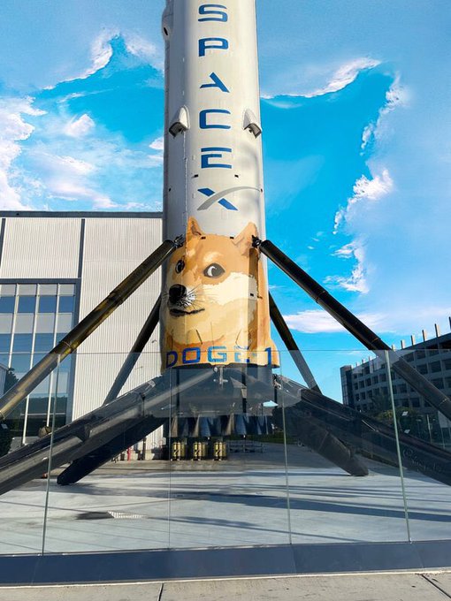A SpaceX Falcon 9 rocket with a Dogecoin logo (Shiba Inu dog) and "DOGE-1" text is displayed on its side, exhibited outside a building.