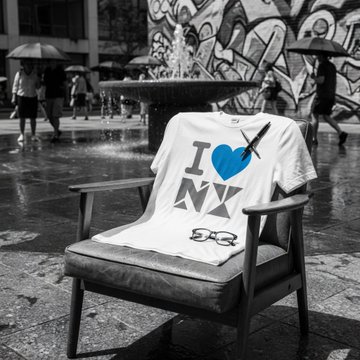 A black-and-white street photograph features a white "I ❤ NY" t-shirt draped over an empty chair in front of a graffiti-covered wall and fountain, with pedestrians carrying umbrellas in the background.