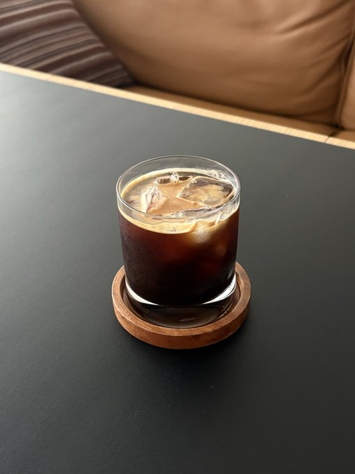 A glass of iced coffee on a wooden coaster sits on a dark table against a backdrop of a brown cushion.