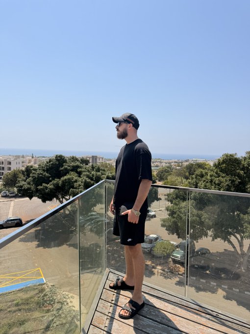 A man in a black cap, t-shirt, and shorts stands on a glass balcony overlooking a coastal city and ocean, capturing a relaxed vacation vibe. Caption: "Posted up where the city meets the sea 🌊☀️"