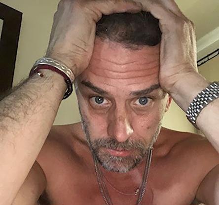 A shirtless middle-aged man with a gray beard stares intensely at the camera with both hands gripping his hair, wearing a watch and a bracelet.