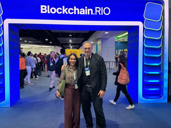 A woman and a man pose together in front of the blue "Blockchain.RIO" entrance at the conference, with attendees visible in the background. The image illustrates the RootstockLabs team's presence at the event, capturing the two team members mentioned (@btccami and @wenciodmartinez) as they engage with the Latin American onchain finance community.